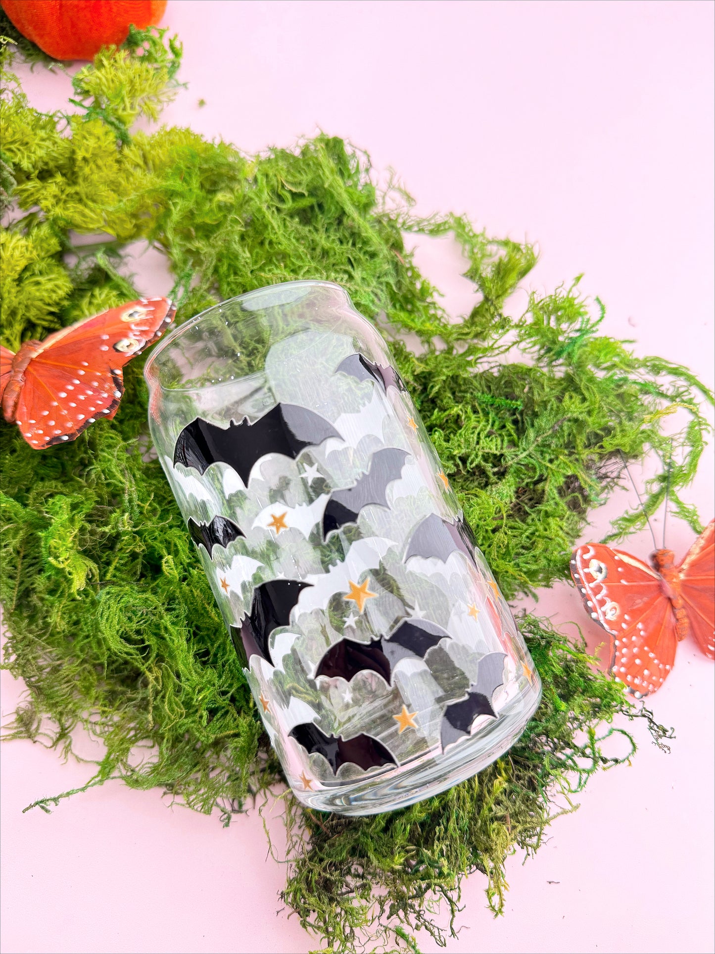 A glass cup with a geometric pattern featuring bats and leaves, displayed on a bed of moss with butterflies and flowers around it.
