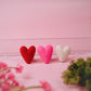 Three heart-shaped objects on a pink surface with blurred flowers and greenery in the foreground.