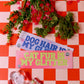 Two signs on a red and white checkered tablecloth with flowers and a small plant.