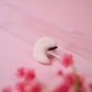 White crescent moon-shaped object on a pink background with blurred pink flowers in the foreground
