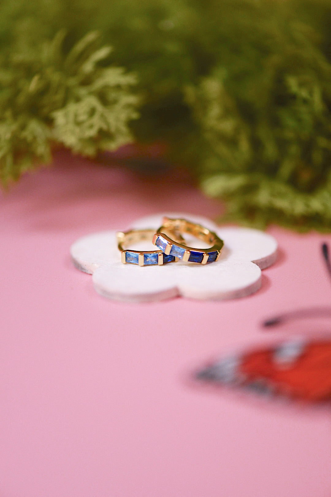 Two gold rings with blue stones on a white flower-shaped object against a pink background with green foliage.