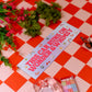 Decorative sign with text on a red and white checkered tablecloth with flowers and a butterfly.