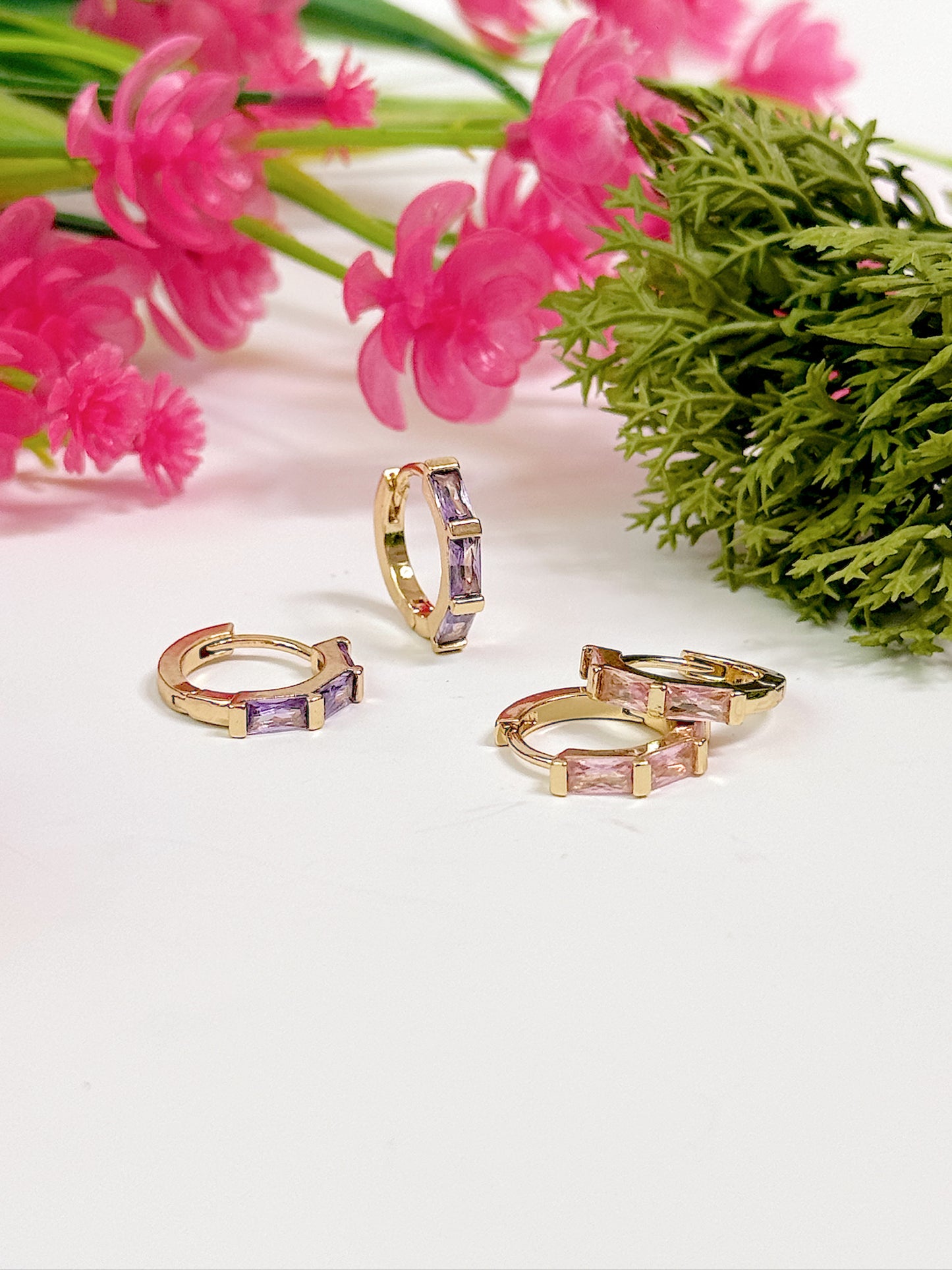Gold hoop earrings with gemstones on a white surface with pink flowers and greenery.