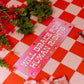 Pink sign with text on a red and white checkered tablecloth with flowers and a glass.
