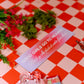 Decorative sign with text on a red and white checkered tablecloth with flowers and a candle.