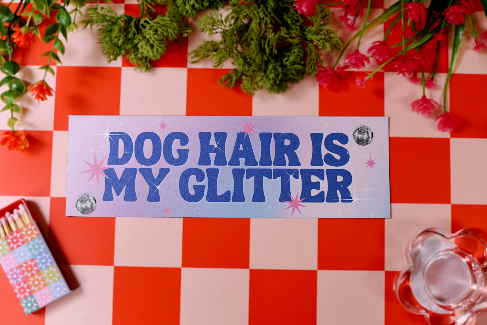 Sign with 'Dog Hair is My Glitter' on a red and white checkered tablecloth with flowers and a matchbox.