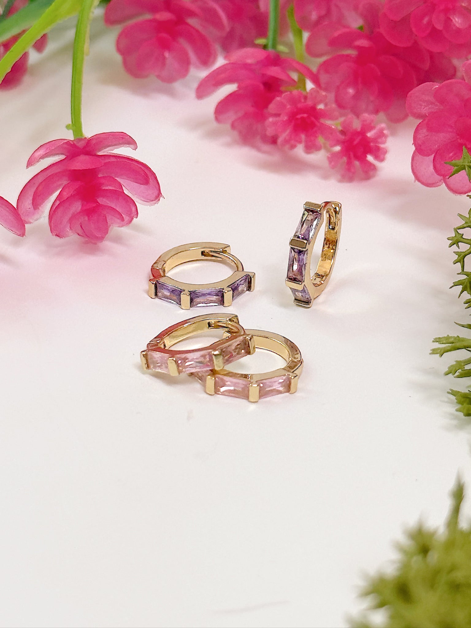 Gold hoop earrings with pink and purple gemstones on a white surface with pink flowers in the background.